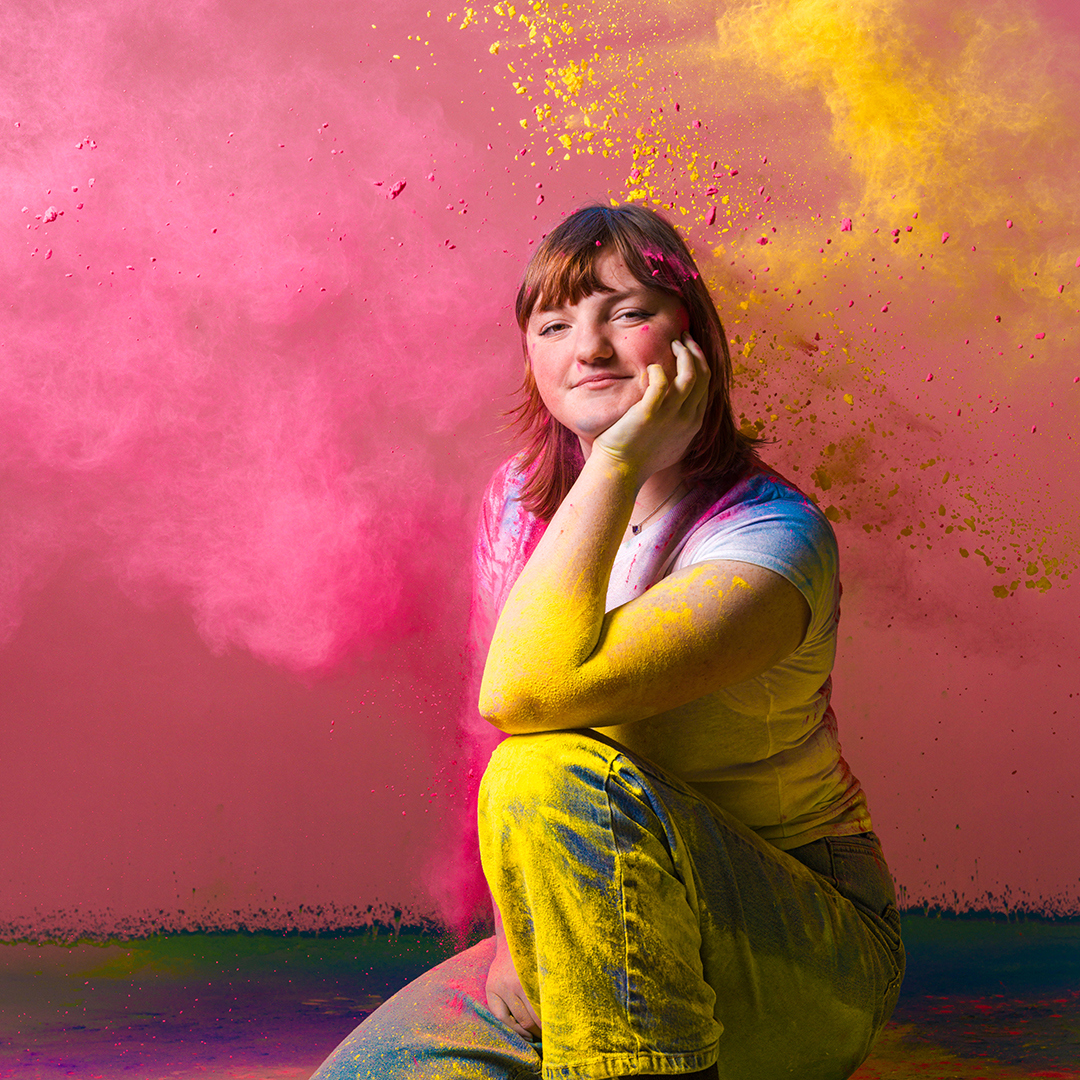 A College age student crouched on the floor on a pink background. She is covered in brightly coloured powder in pink and yellow.