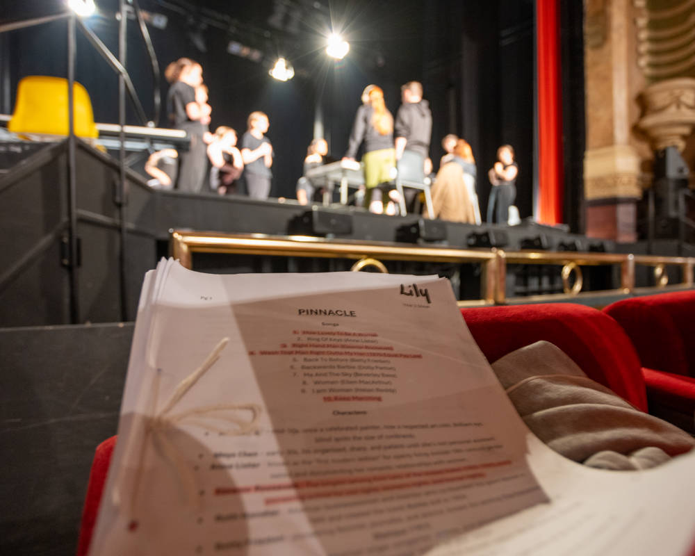 The script of Pinnacle in the foreground with students rehearsing in the background.