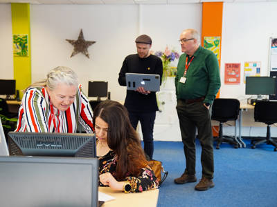Staff supporting a student using a computer while colleagues work in the background.