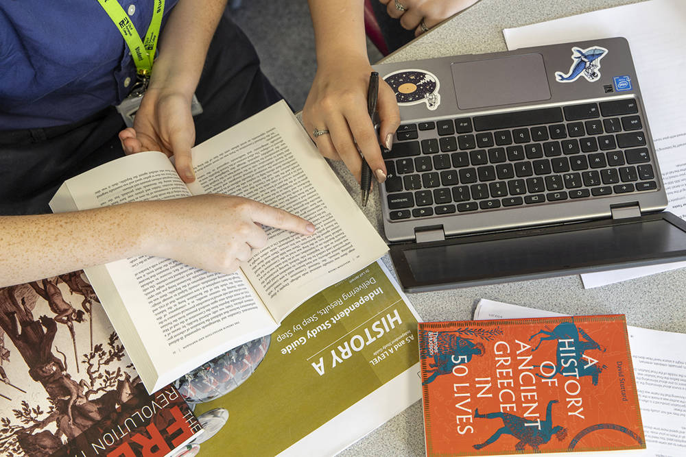 A birds-eye-view of a table containing books, a laptop and other study notes as two students study collaboratively.