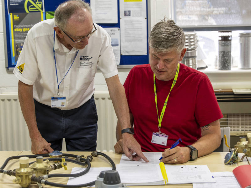 A college tutor supporting an adult learner during a gas training session, reviewing paperwork together at a workbench with gas equipment.