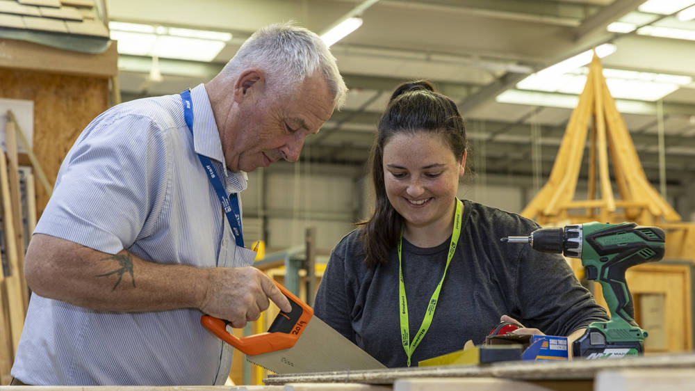 Carpentry teacher sawing wood in the workshop at North Harbour Campus with a student watching
