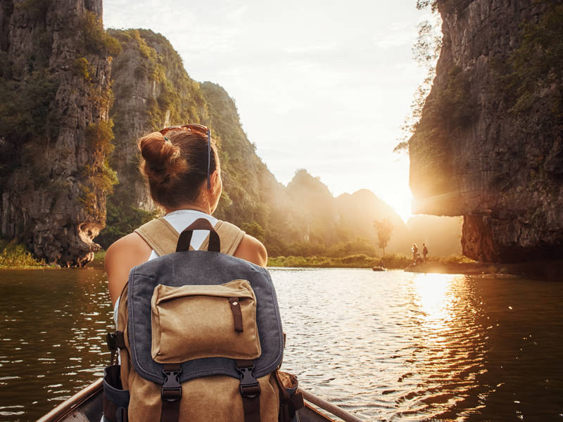 Woman with backpack swims on boat among karst mountains to meet her friends