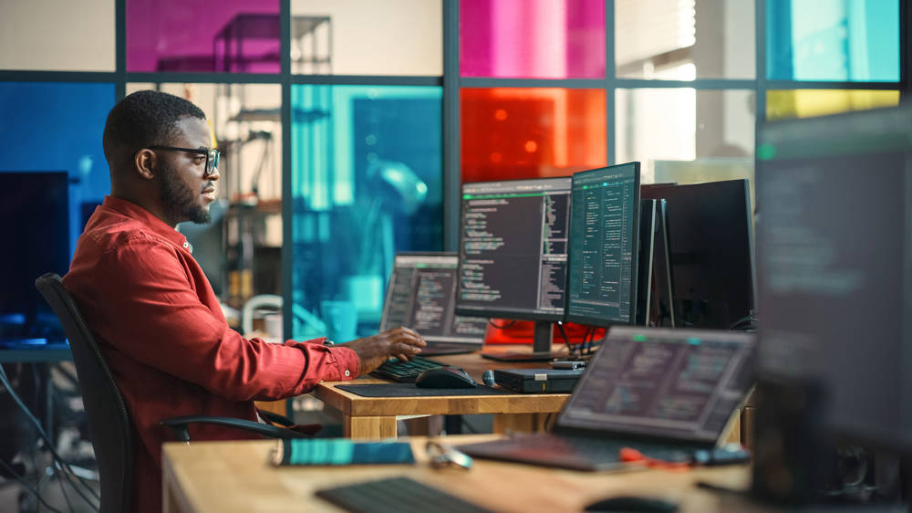 African American man on desktop computer with multiple monitors and laptop in creative office