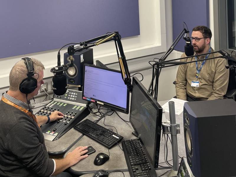 COPC Assistant Principal Lee McCarthy sits facing the camera at a DJ booth at Express FM, interviewed by Ian James.