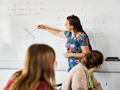 Maths teacher explaining a graph and equations on a whiteboard during a classroom lesson.