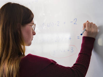 Student writing maths calculations and equations on a classroom whiteboard.