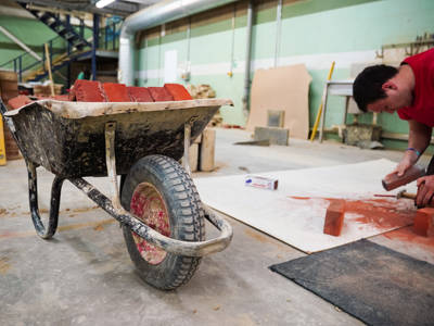 A wheelbarrow of bricks and a student working in the bricklaying workshop 