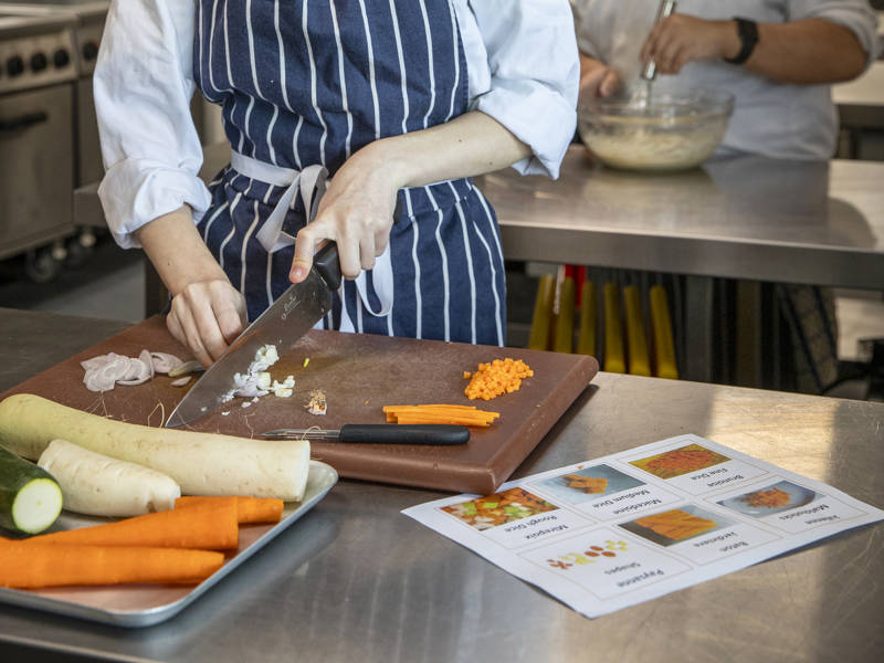Catering student preparing vegetables in a professional training kitchen while other students work in the background.