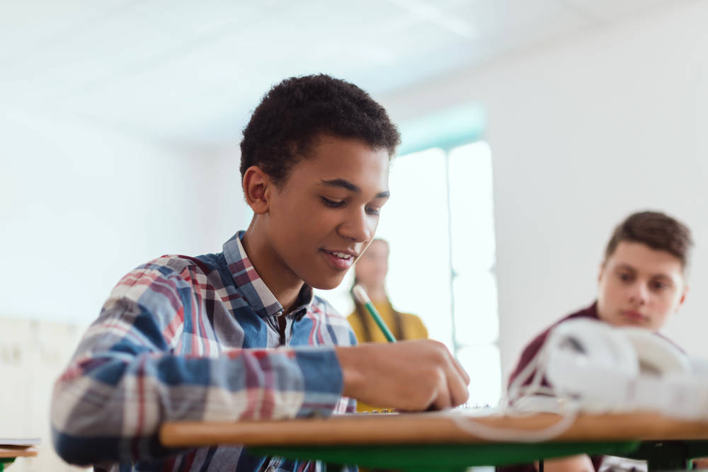african american teenage school student writing in textbook and classmates behind