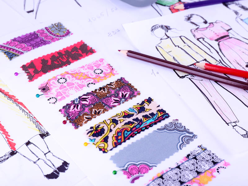 An image of pinned fabric swatches and fashion drawings on a table with coloured pencils and measurement notes.