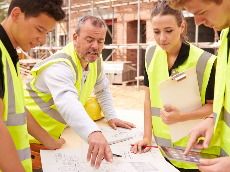 Builder On Building Site Discussing Work With Apprentice