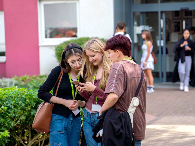 Three students looking at a friends phone outside the entrance of the Sixth Form Campus
