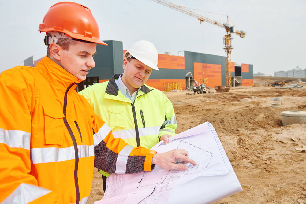 Two men in hard hats and hi-vis jackets looking at plans on a construction site.